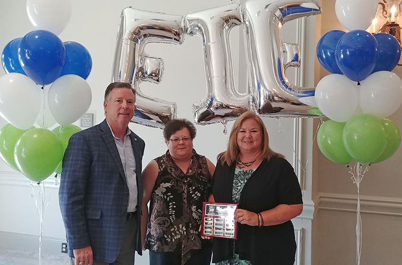 Three people in business dress surrounded by green, white and blue and silver balloons, one of them holding a plaque.