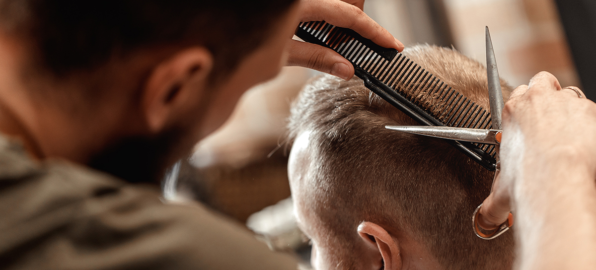 Man with a full ebeard trimming hair of another man with scissors and comb