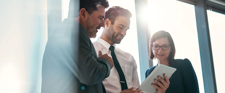 Three office employees of various experience collaborate together with a mobile device by a large window.