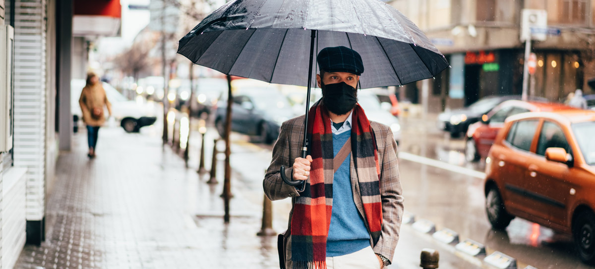 Caucasian man in blazer, scarf and black facemask holding an open grey umbrella as he walks down a city street.